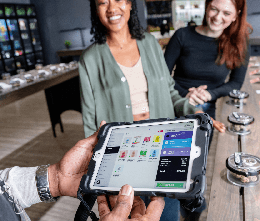A budtender holds a tablet displaying Meadow’s POS system while assisting two smiling customers at a cannabis dispensary. The budtender is not behind a counter, illustrating the floating sales model where staff can move freely throughout the store to provide a more personalized, flexible shopping experience.