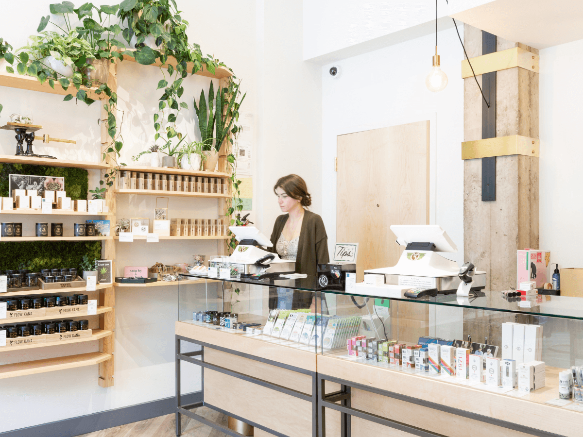 The image depicts a vibrant and welcoming scene inside the Vapor Room, a premier San Francisco dispensary with a rich history dating back to 2004. The dispensary's interior is bright and filled with natural light, creating an inviting atmosphere. A sales associate stands behind the counter, engaging with a customer while efficiently using Meadow's point-of-sale system. The background showcases neatly organized shelves filled with an array of cannabis products, highlighting the dispensary's commitment to excellence in product selection. The overall ambiance reflects the unique culture and legendary community spirit that the Vapor Room has cultivated over the years.