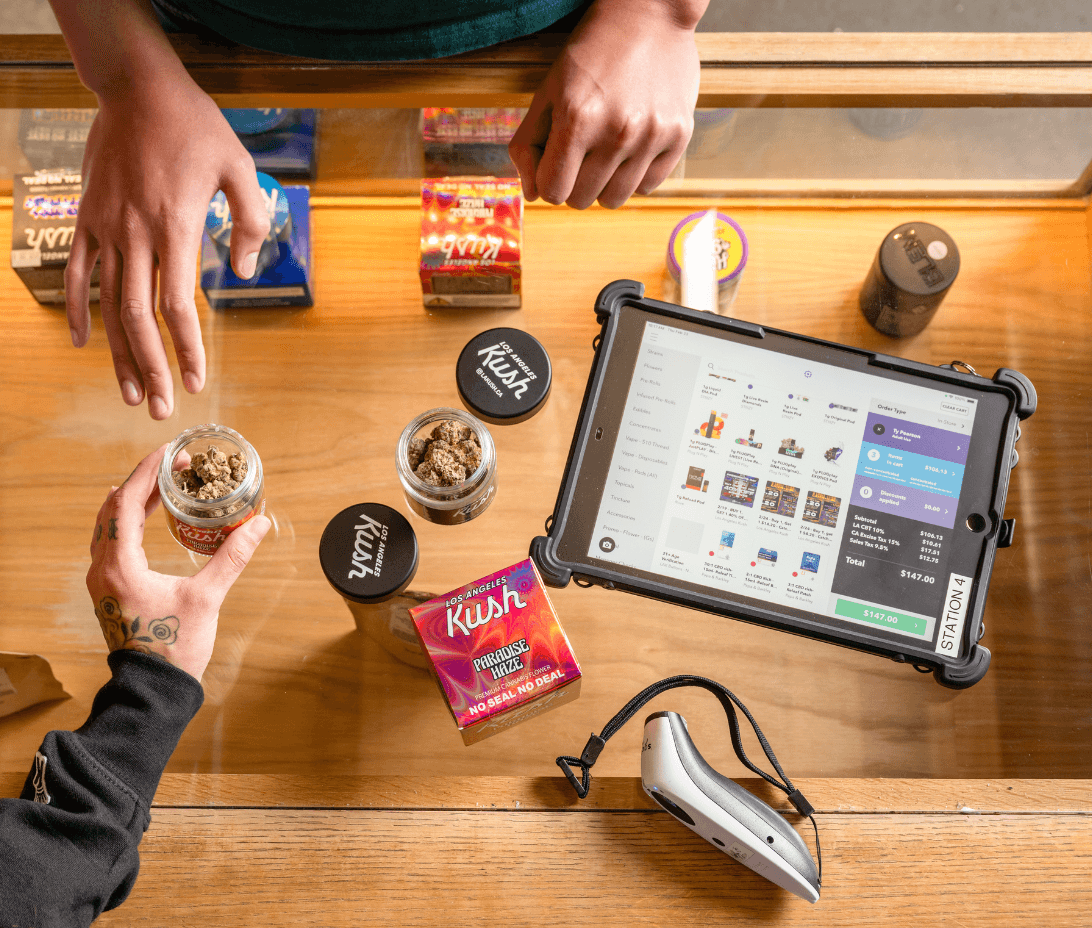 A budtender and customer interact at a glass display counter stocked with premium cannabis flower and packaged products. Meadow’s point-of-sale system is shown on a tablet, with a clear interface displaying the customer’s order summary and total. A barcode scanner rests on the counter nearby. This image highlights a modern, efficient checkout experience in a California dispensary, powered by Meadow’s intuitive cannabis retail software.