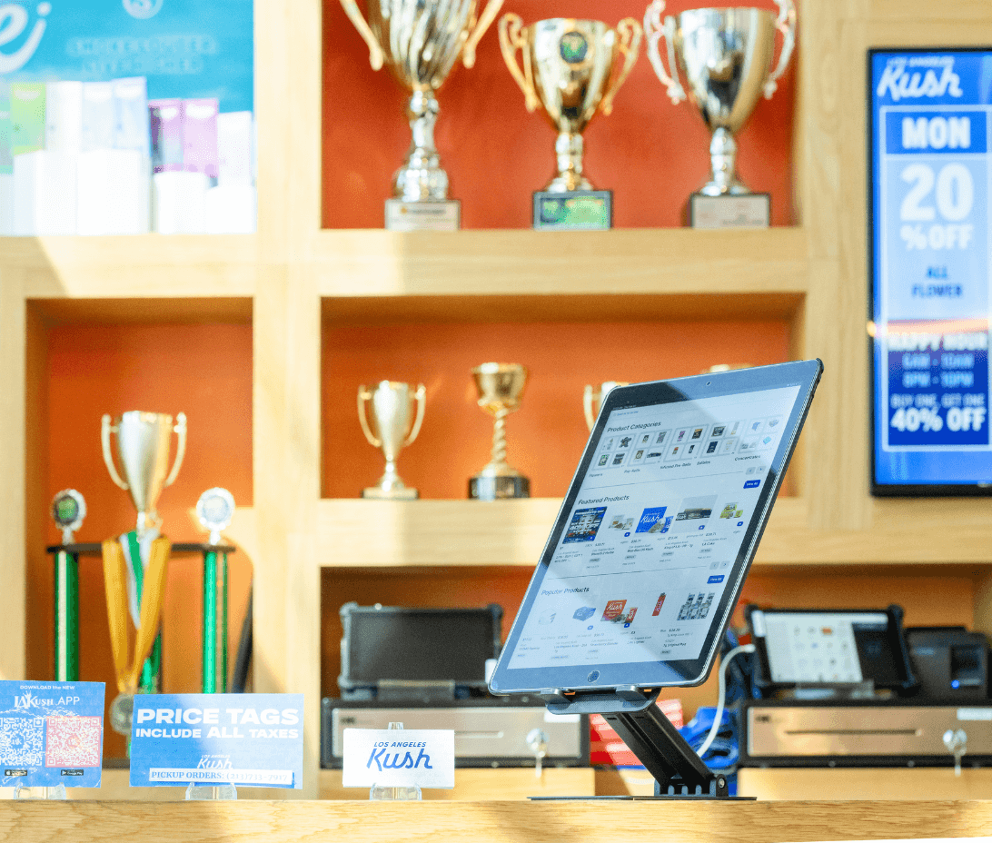 A self-service kiosk running Meadow’s eCommerce platform sits on the counter of a bright and modern California dispensary, enabling customers to browse and place pickup orders with ease. Behind the kiosk, a wall of trophies highlights the shop’s award-winning status, while signage promotes daily deals and tax-inclusive pricing. This image illustrates how Meadow supports fast, compliant retail transactions and customer autonomy—ideal for California’s competitive, high-volume cannabis market.