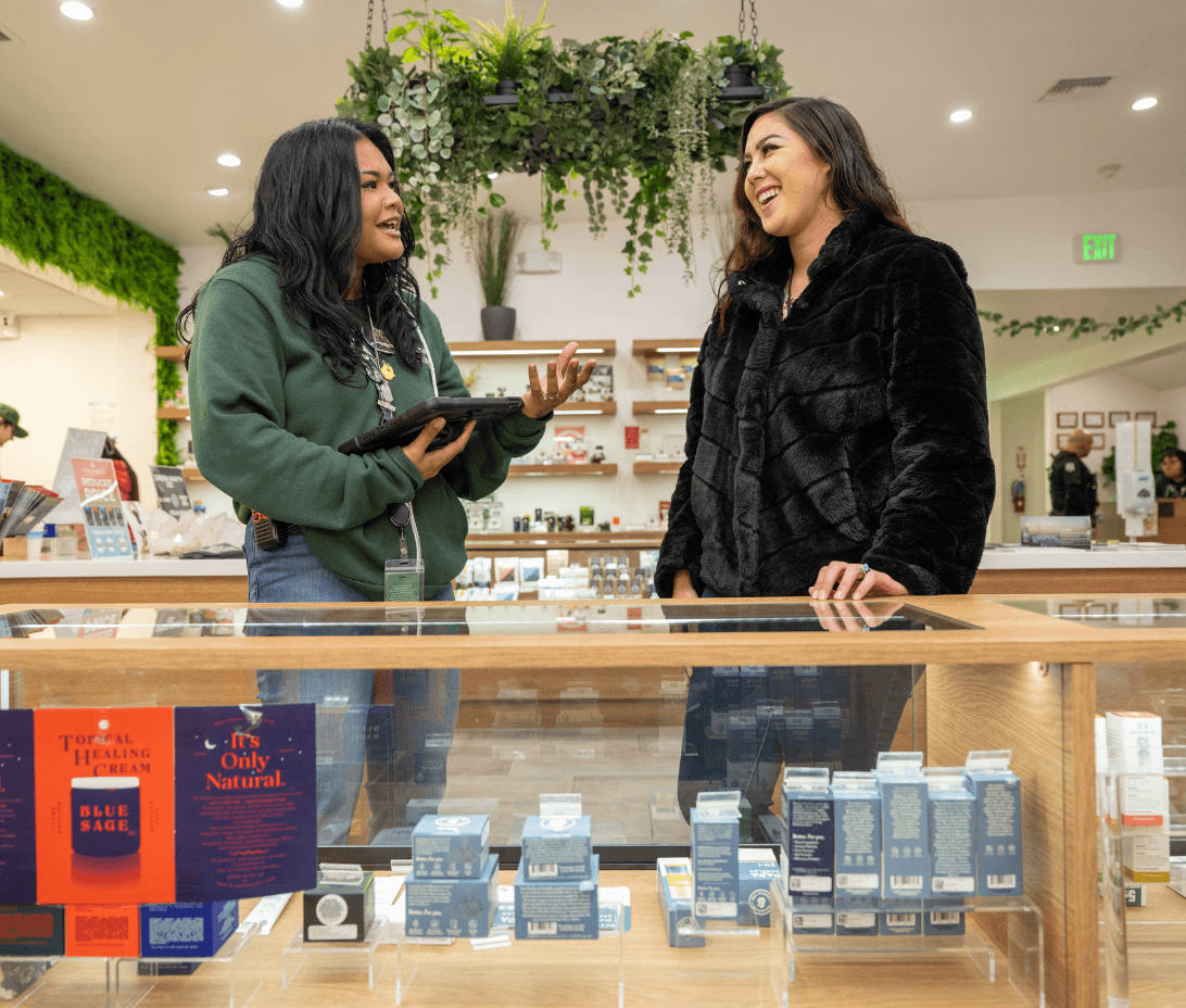Inside a modern, inviting dispensary, a knowledgeable budtender is assisting a customer with a smile while showing options on a digital tablet and pointing out specific cannabis products that suit the customer's preferences. The counter displays an array of cannabis products under soft, inviting lighting, creating a comfortable and personalized shopping experience. This scene illustrates Meadow's commitment to exceptional customer service, highlighting the importance of expert advice, quick issue resolution, and personalized care in becoming a favorite dispensary.