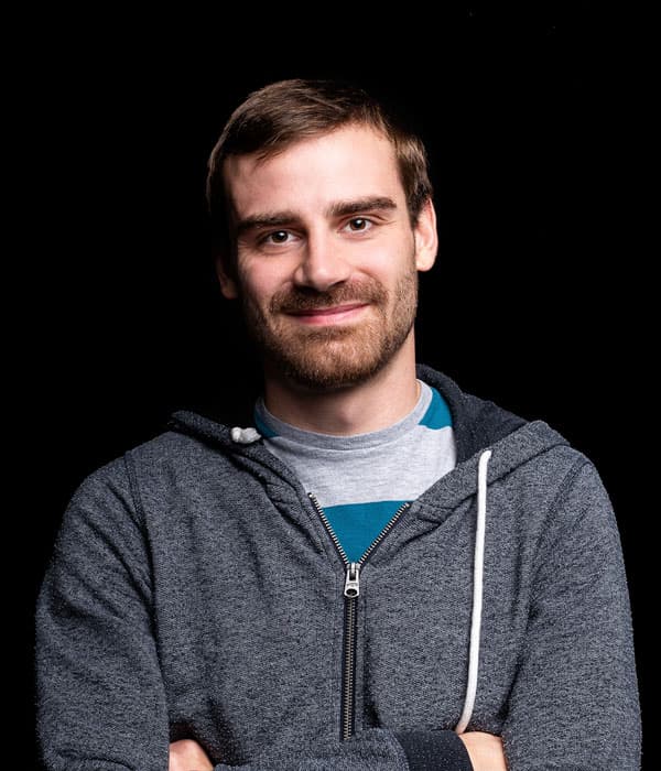 A slightly smiling white man with brown hair and a brown beard, wearing a gray hoodie over a light colored shirt on a black background.