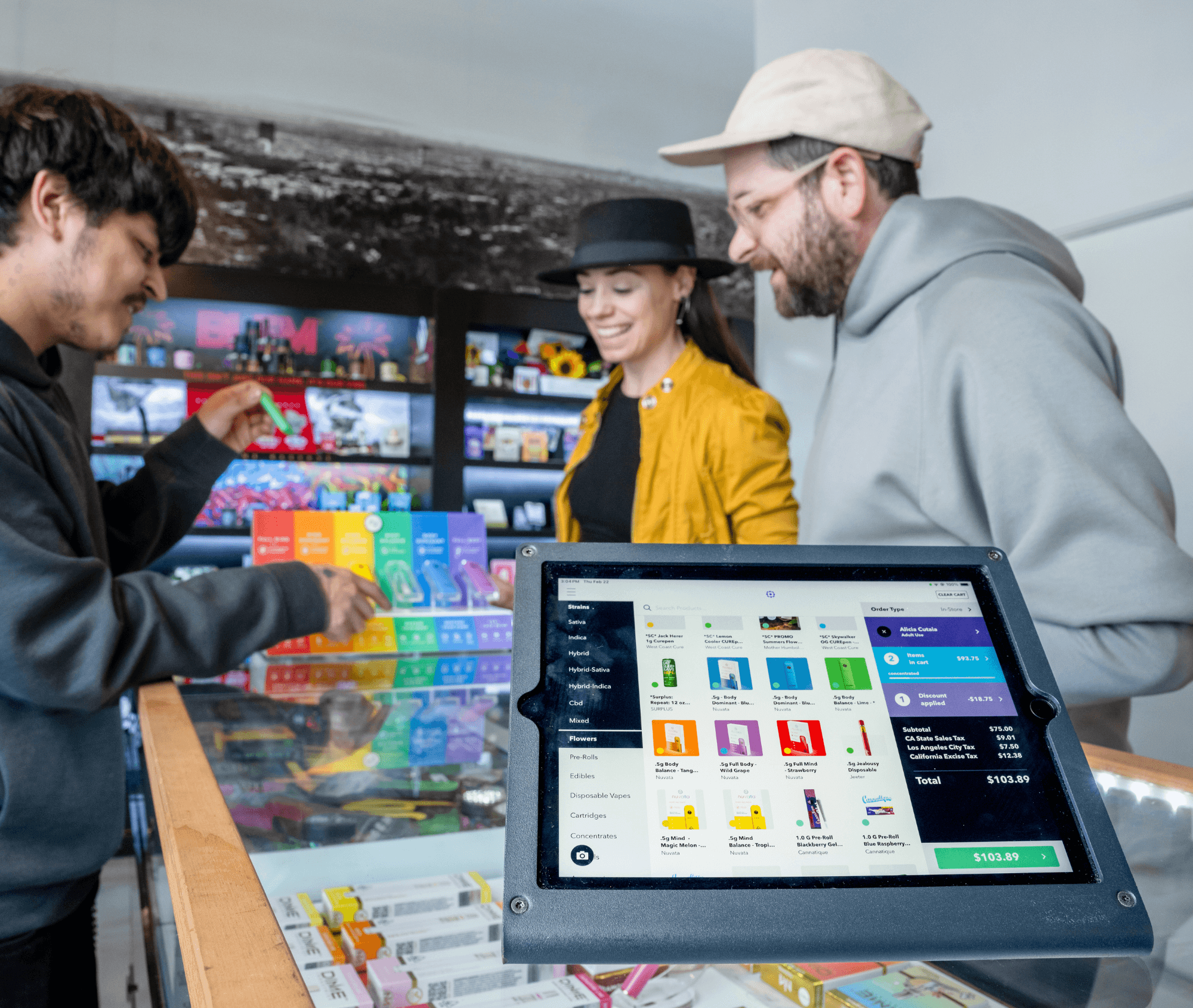 A close-up of a tablet displaying a cannabis point-of-sale screen sits atop a dispensary counter as two customers happily explore colorful cannabis product boxes with a budtender. The interface shows live product availability and checkout details.