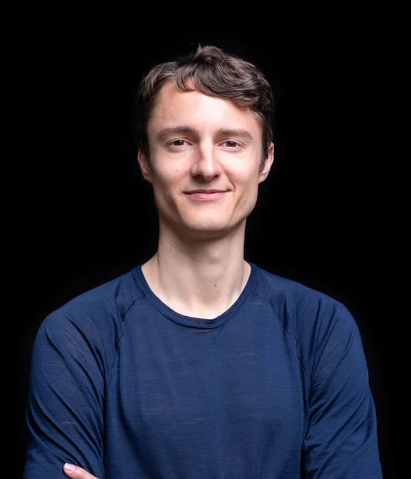 A white man with short brown hair, smiling slightly, wearing a brown long sleeve shirt, on a black background.