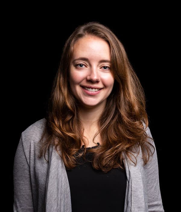 A young white woman with shoulder length brow hair, smiling, wearing a gray cardigan, on a black background.