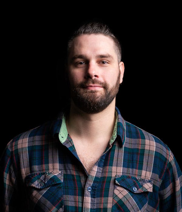 A white man with slicked back brown hair and a beard, wearing a blue flannel shirt, on a black background.