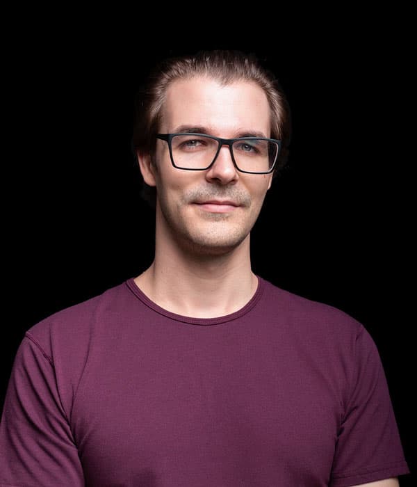 A slightly smiling white man with slicked back brown hair and glasses, wearing a wine-colored shirt in front of a black backdrop.