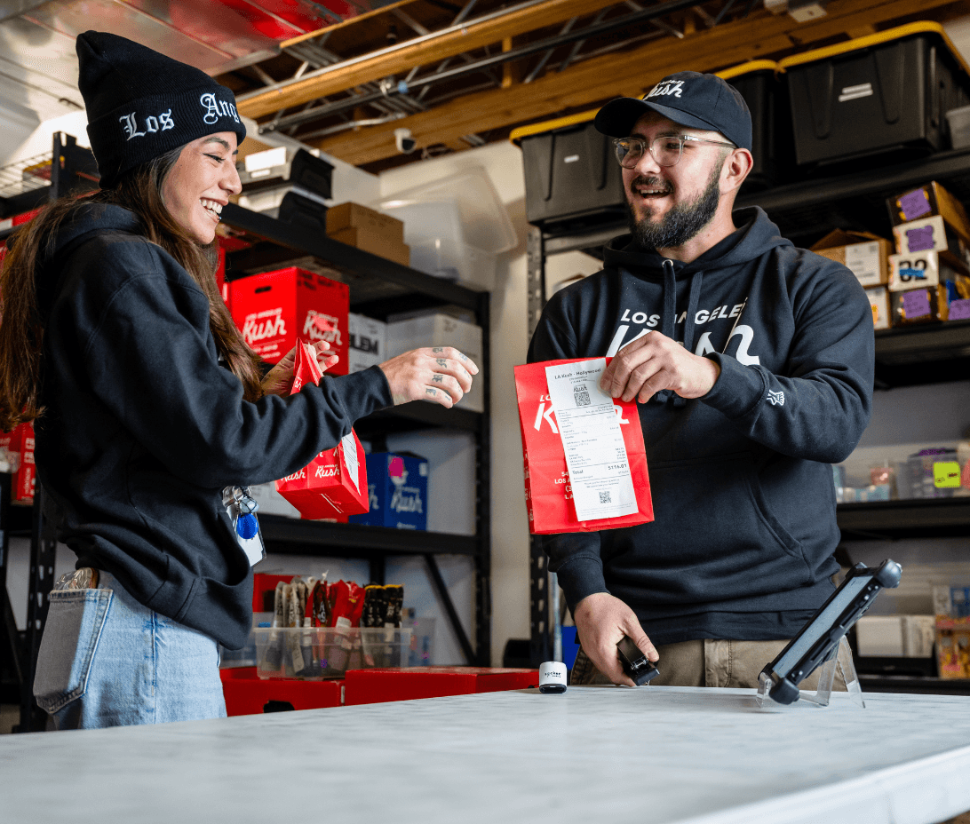 Two dispensary team members laugh and work together in a cannabis fulfillment area, preparing an order with branded LA Kush packaging. One holds a barcode scanner while the other applies the printed receipt, using Meadow’s fulfillment tools to manage accurate, compliant order prep. Organized bins and labeled shelves reflect an efficient operation. This image captures the collaborative, fast-paced energy of California cannabis retail backed by Meadow’s end-to-end fulfillment workflows.