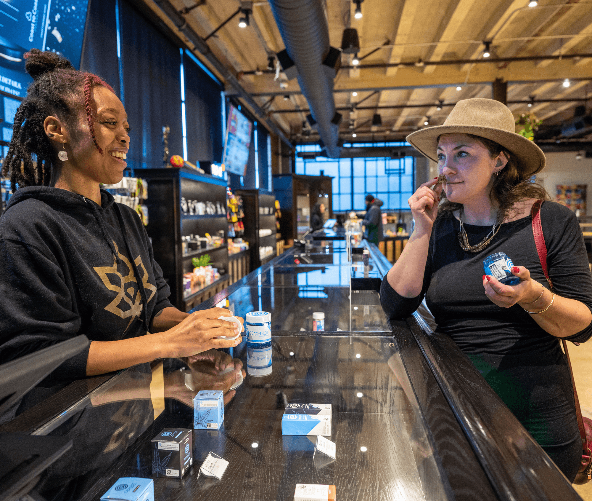 A smiling budtender chats with a customer who is smelling a cannabis bud from a jar, both standing at a glass display counter filled with cannabis products. The dispensary has a warm, inviting interior with wood-beam ceilings and modern lighting.