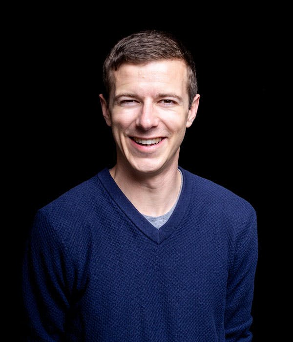 A smiling white man with brown hair wearing a blue v-neck shirt on a black background.